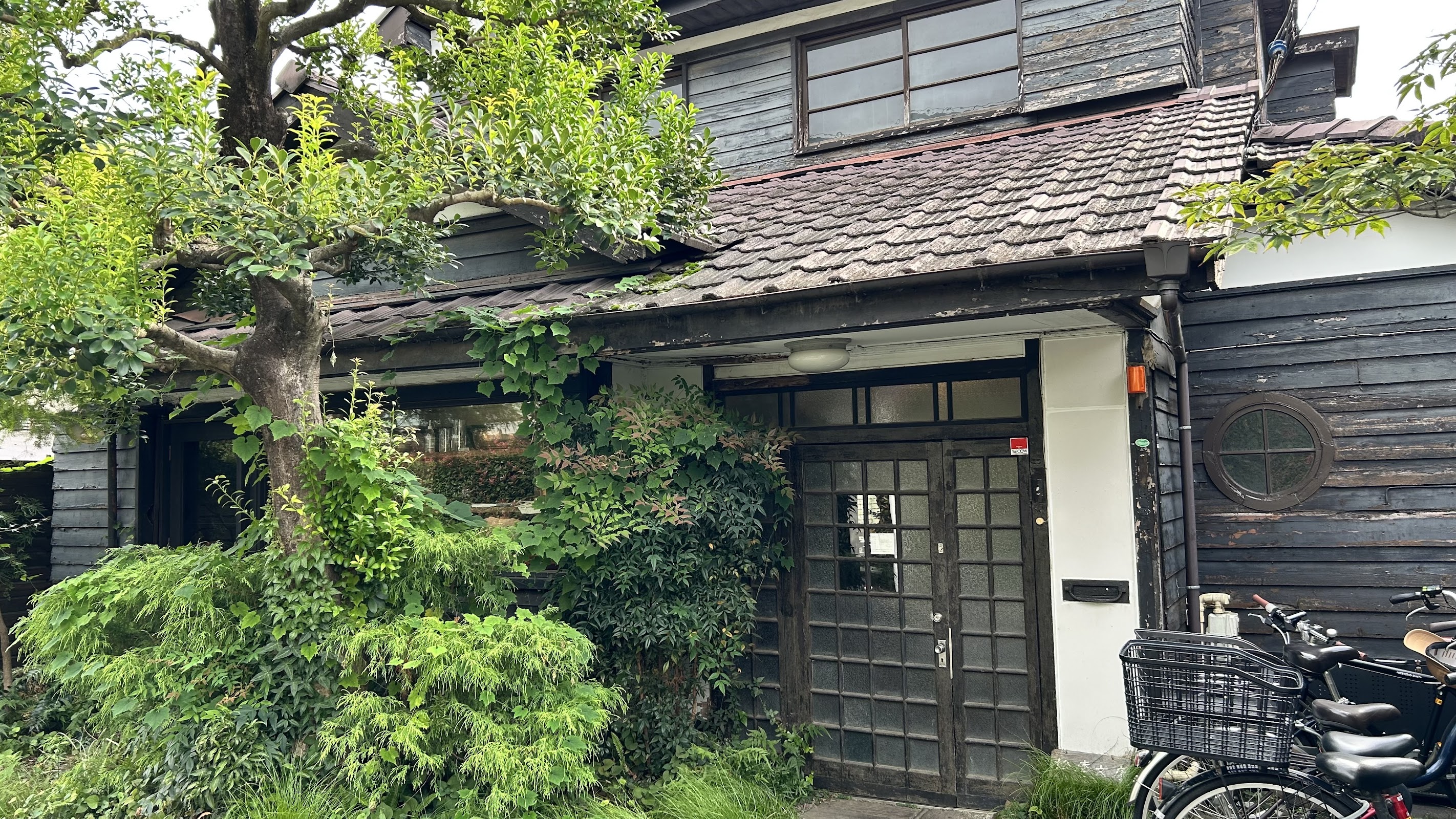 緑に囲まれた古民家の正面玄関。瓦屋根に苔がのり、生垣の脇に自転車が置かれている。